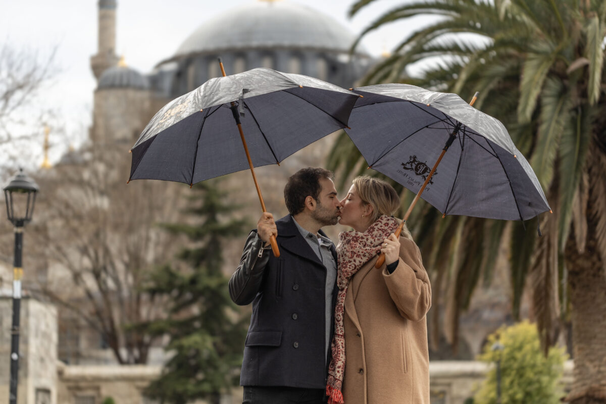 A couple shares a kiss under two separate umbrellas in front of the Blue Mosque in Istanbul, surrounded by palm trees and a cloudy winter sky.