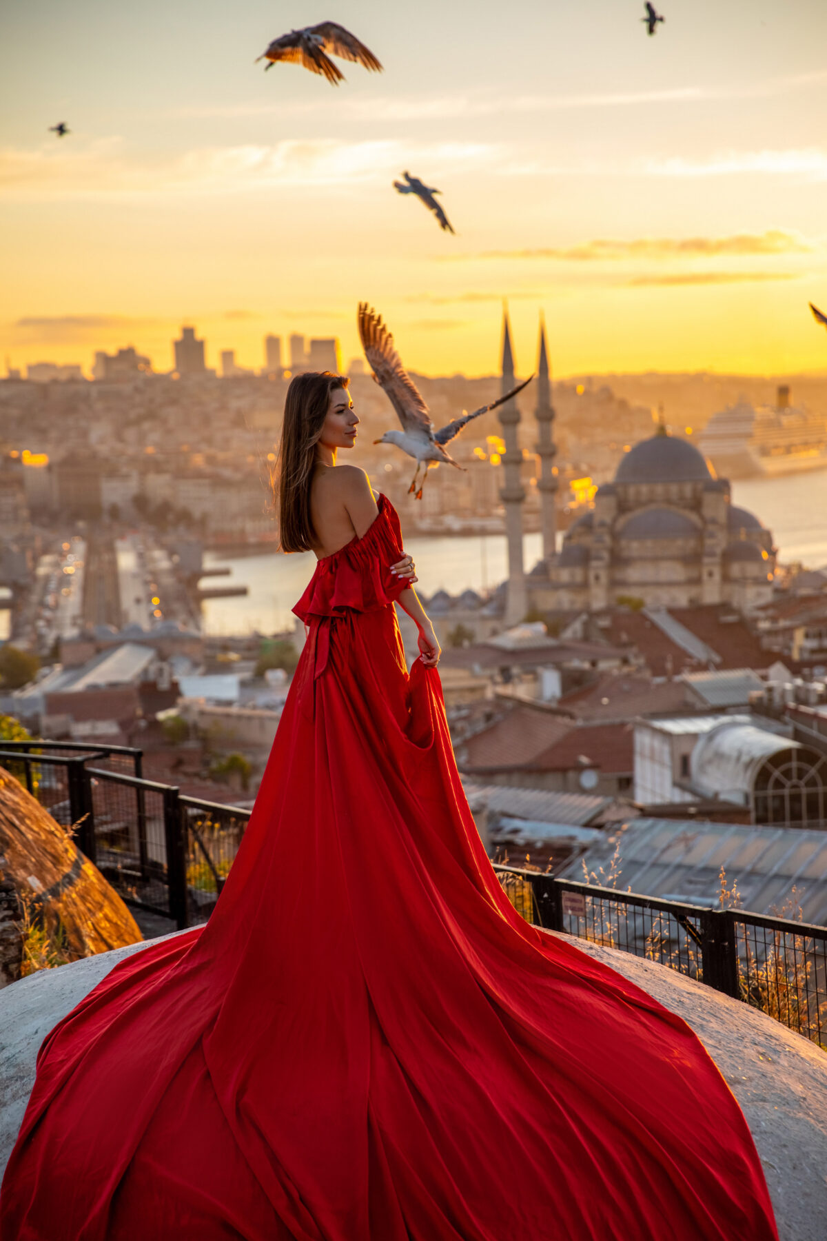 Woman in a flowing red dress watching the sunset over Istanbul with birds flying and a historic mosque in the background